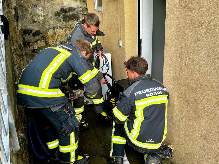 Vier Feuerwehrleute in Uniformen arbeiten an einer Aufgabe in einem engen Raum, wahrscheinlich am Eingang eines Hauses. Einer hält einen Schlauch, ein anderer eine Kamera, und zwei beobachten.