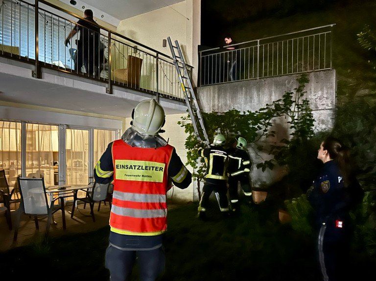 Feuerwehrleute stehen vor einem Apartmentgebäude. Eine Person ist auf dem Balkon, andere sind am Boden. Eine Leiter lehnt an der Wand.