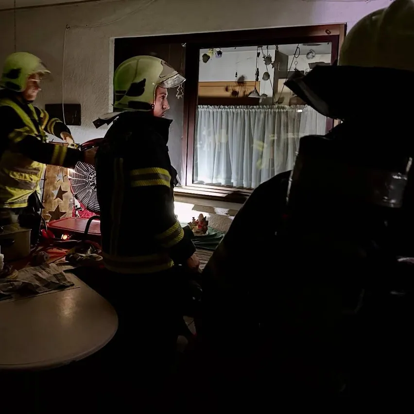 Drei Feuerwehrleute in Schutzausrüstung stehen in einem Raum. Einer von ihnen hat einen Ventilator in der Hand. Es gibt einen Tisch mit Objekten darauf.