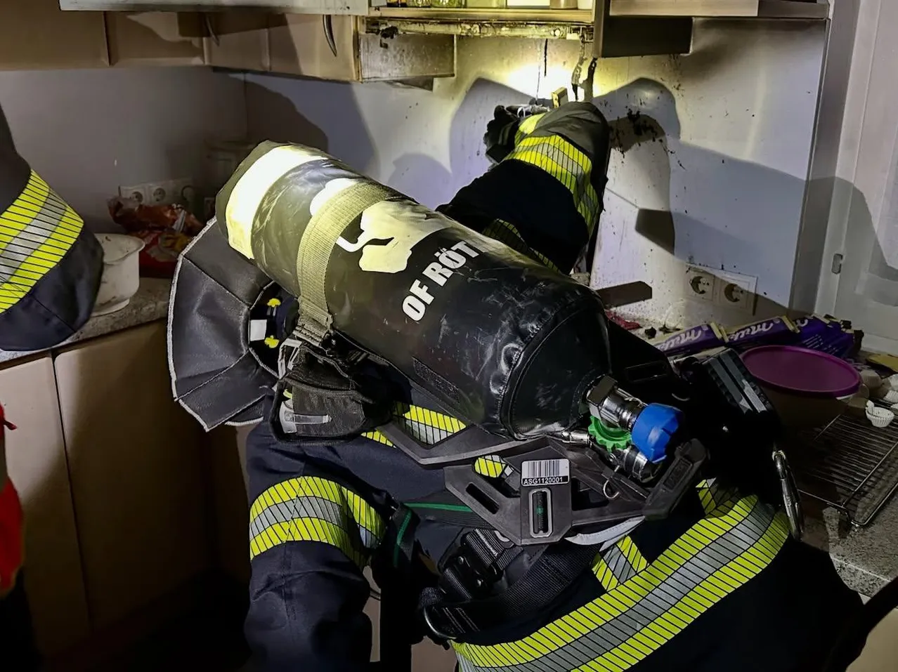 Ein Feuerwehrmann in einem gelben und schwarzen Anzug arbeitet in einem Raum, trägt eine Maske und trägt eine Gasflasche.