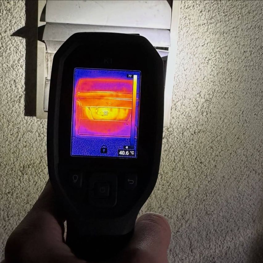 Eine Person hält ein schwarzes Gerät, das ein Wärmebild einer Wand zeigt und eine Temperatur von 40,6 Grad Celsius anzeigt.