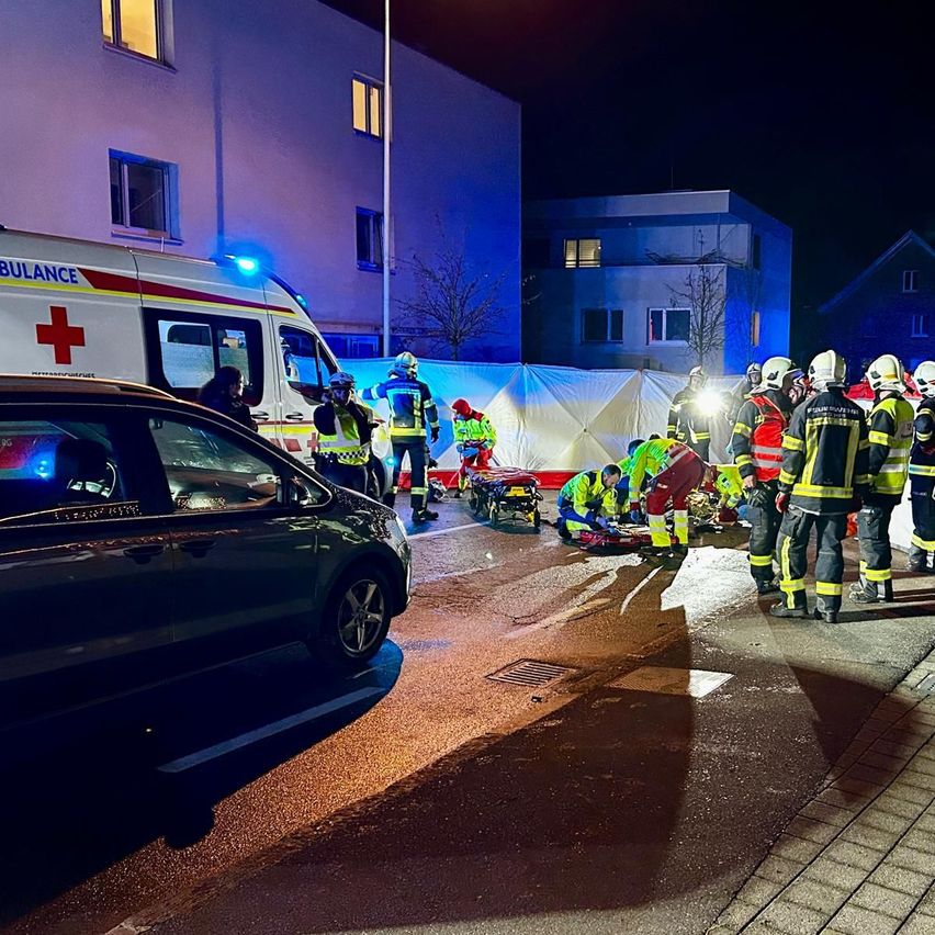Rettungskräfte kümmern sich nachts auf einer Straße um einen Unfall, wobei ein Krankenwagen und Feuerwehrleute anwesend sind. Im Hintergrund sind Gebäude und ein Auto zu sehen.