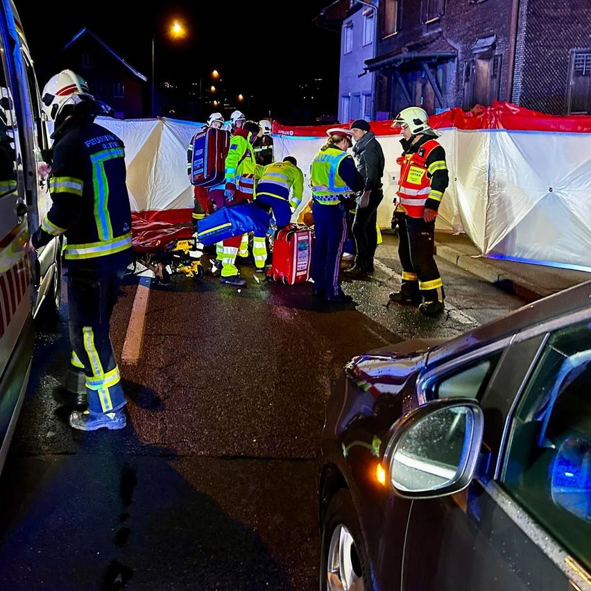 Eine Gruppe von Rettungskräften kümmert sich um eine verletzte Person auf der Straße. Ein Krankenwagen ist in der Nähe geparkt, und ein Auto ist ebenfalls vorhanden.