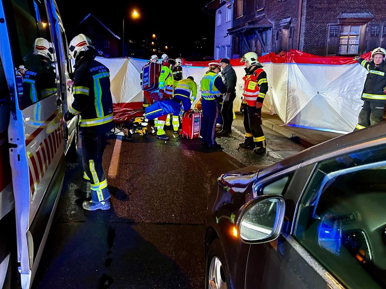 Eine Gruppe von Rettungskräften kümmert sich um eine verletzte Person auf der Straße. Ein Krankenwagen ist in der Nähe geparkt, und ein Auto ist ebenfalls vorhanden.