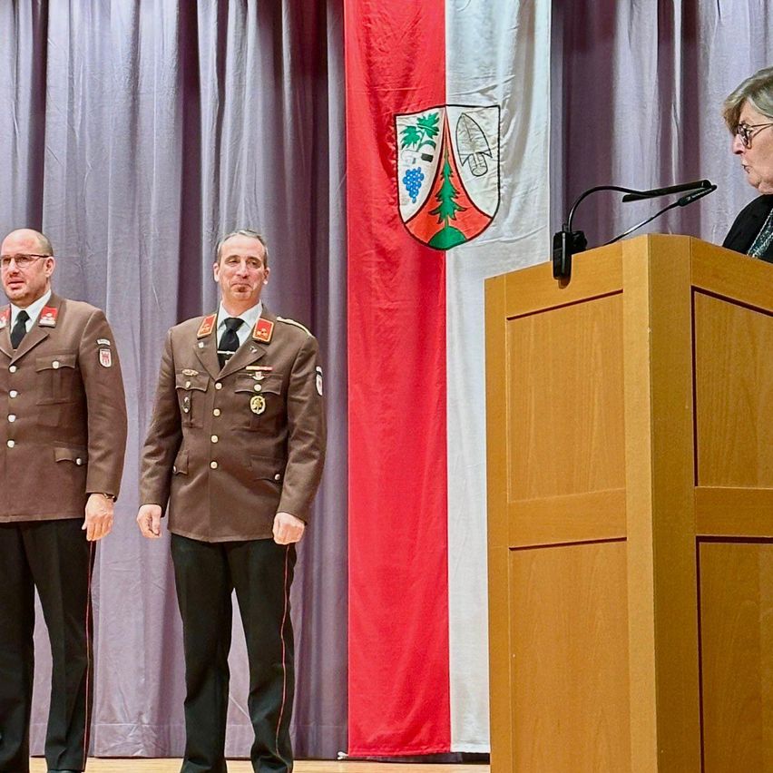 Zwei Militäroffiziere in braunen Uniformen stehen nebeneinander auf der Bühne. Eine Frau spricht in ein Mikrofon an einem Podium. Dahinter hängt eine Flagge mit einem Wappen.