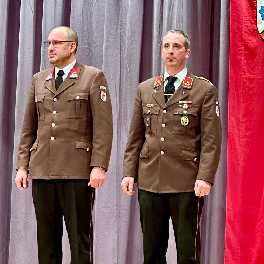 Zwei Männer in Militäruniformen stehen nebeneinander und schauen nach vorne. Beide tragen eine Brille und eine Krawatte, und ihre Uniformen sind mit Medaillen und Abzeichen geschmückt. Hinter ihnen ist eine rote Flagge und graue Vorhänge zu sehen.