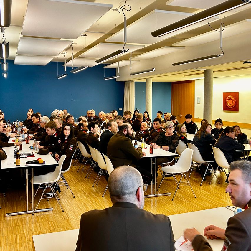 Eine Gruppe von uniformierten Personen sitzt in einem Konferenzraum mit blauen Wänden und Holzböden an Tischen. Flaschen und Teller stehen auf den Tischen.