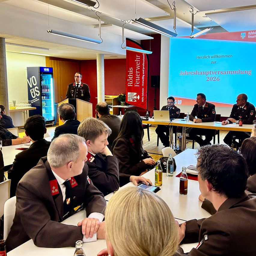 Eine formelle Versammlung in einem Konferenzraum. Ein Mann in Uniform steht an einem Podium und spricht eine Zuhörerschaft an. Die Leinwand hinter ihm zeigt eine Präsentation mit dem Titel Jahreshauptversammlung 2026.