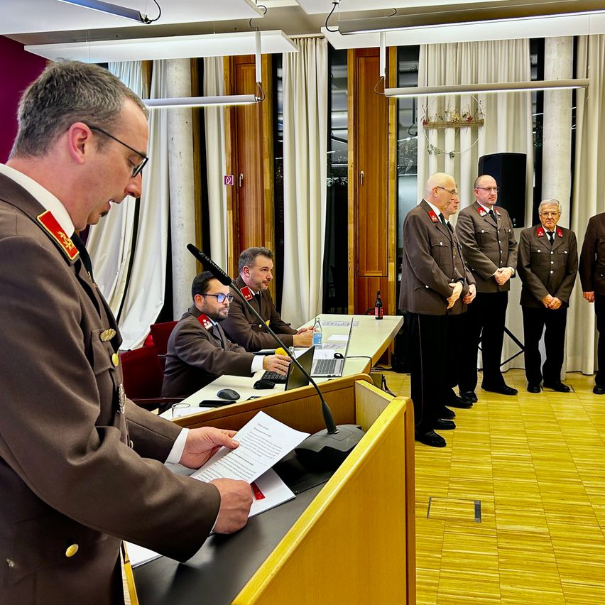 Ein Mann in Uniform steht an einem Podium, liest ein Papier, während andere in Uniform hinter ihm stehen. Ein Schreibtisch mit Laptop und anderen Gegenständen steht hinter dem Podium.