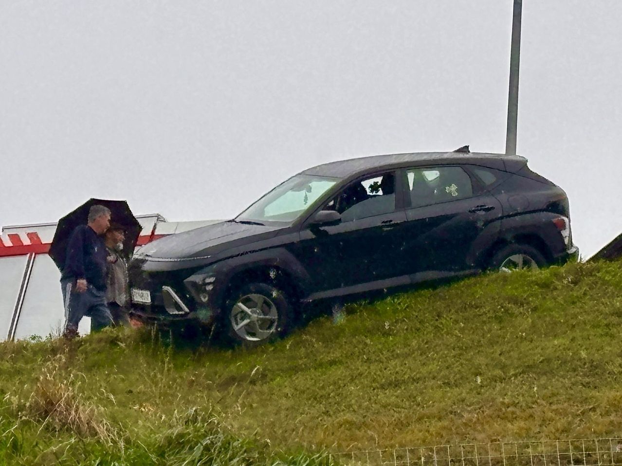 Ein schwarzer SUV steckt auf einem Grashügel fest, ein Mann mit einem Regenschirm steht in der Nähe. Der SUV scheint gerade die Straße verlassen zu haben.