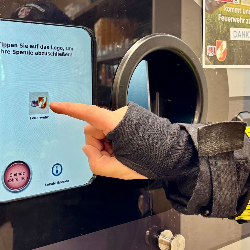 Eine Hand zeigt auf einen Spendenbildschirm an einer Spendenmaschine, mit einem Button, der 'Spende abbrechen' liest und einem Informationssymbol daneben. Hinter dem Bildschirm zeigt ein Plakat 'DANK' und ein Feuerwehremblem.