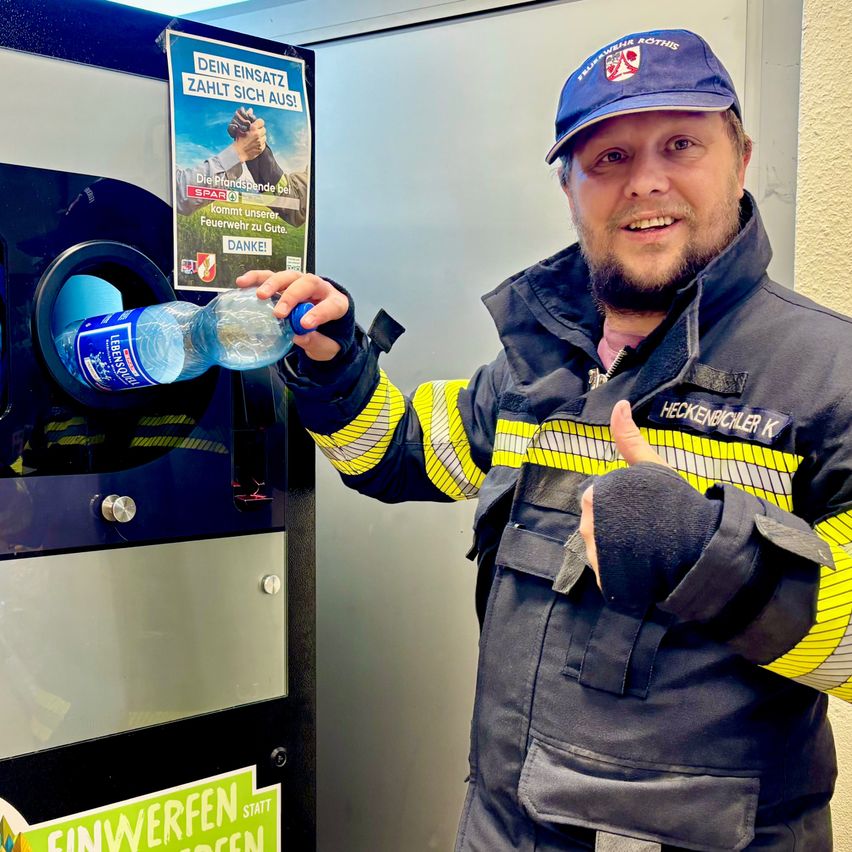 Ein Mann in einer Feuerwehruniform hält eine Wasserflasche, lächelt und gibt einen Daumen hoch vor einem Recyclingbehälter mit einem Schild, das 'Ihre Unterstützung zählt!' steht.