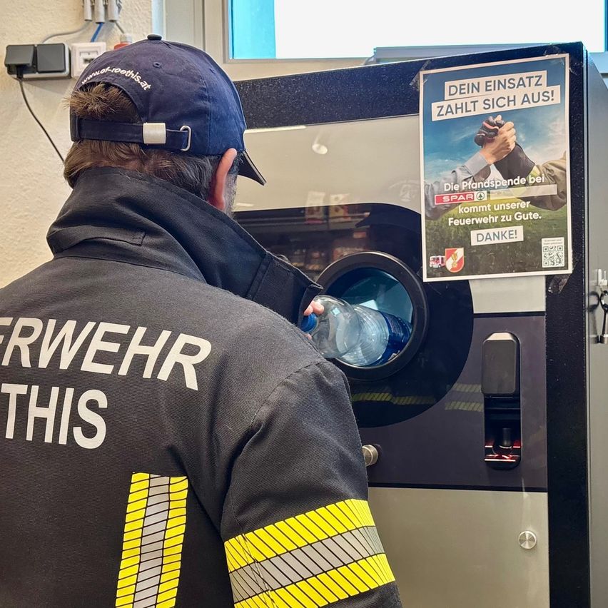 Ein Feuerwehrmann füllt eine Flasche an einer Wasser-Nachfüllstation. Die Station hat ein Plakat, das 'Dein Einsatz zahlt sich aus!' besagt.