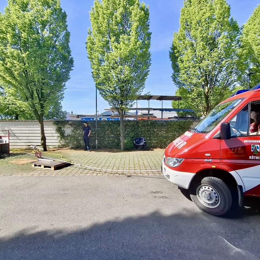 Ein roter und weißer Van ist auf einem Parkplatz geparkt, mit einem Mann hinter einem Busch. Bäume und ein Gebäude sind im Hintergrund.