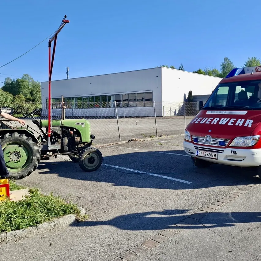 Ein grüner Traktor ist neben einem weißen Gebäude und einem roten Feuerwehrauto geparkt. Eine Person steht neben dem Traktor. Bäume und Pflanzen sind im Hintergrund zu sehen.