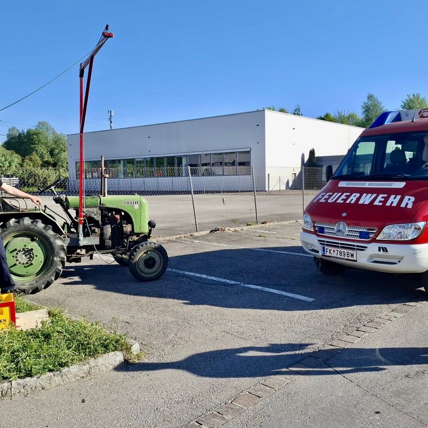 Ein grüner Traktor ist neben einem weißen Gebäude und einem roten Feuerwehrauto geparkt. Eine Person steht neben dem Traktor. Bäume und Pflanzen sind im Hintergrund zu sehen.