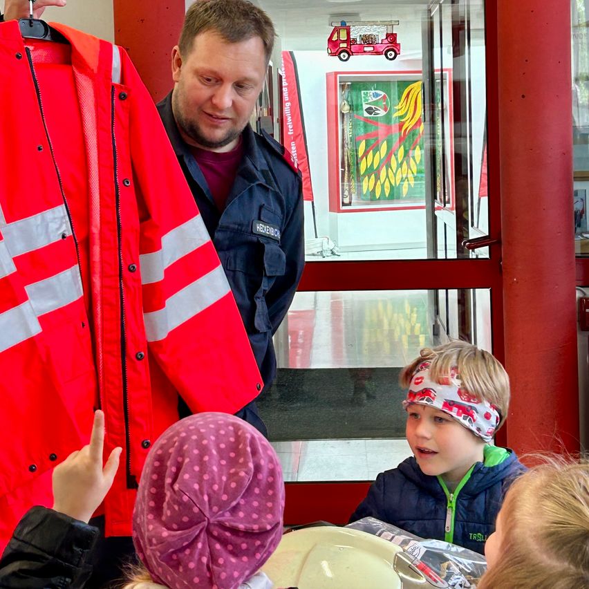 Ein Mann in Uniform zeigt Kindern eine rote Sicherheitsjacke. Sie sitzen um einen Tisch mit Tellern und scheinen aufgeregt zu sein.