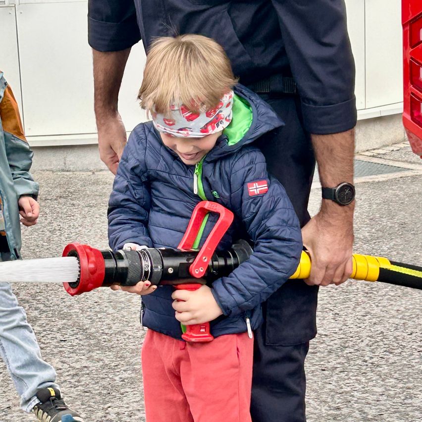 Ein junger Junge hält einen rot-schwarzen Feuerwehrschlauch, geführt von einem Feuerwehrmann. Hinter ihm steht ein weiteres Kind. Sie sind draußen auf einer Betonfläche.