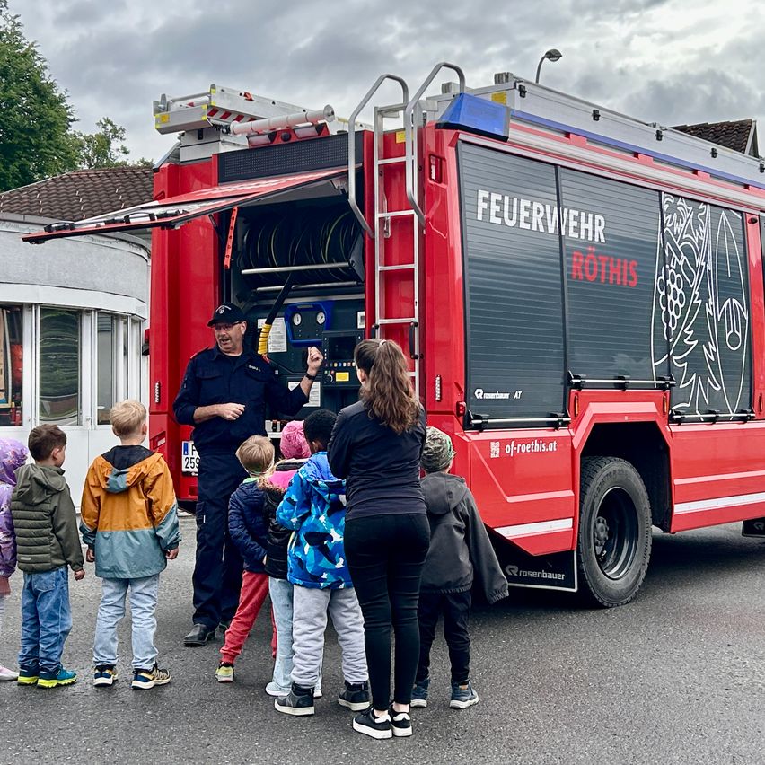 Ein Feuerwehrmann spricht vor einem roten Feuerwehrauto mit Kindern. Das Auto ist mit 'Feuerwehr Rothis' beschriftet.
