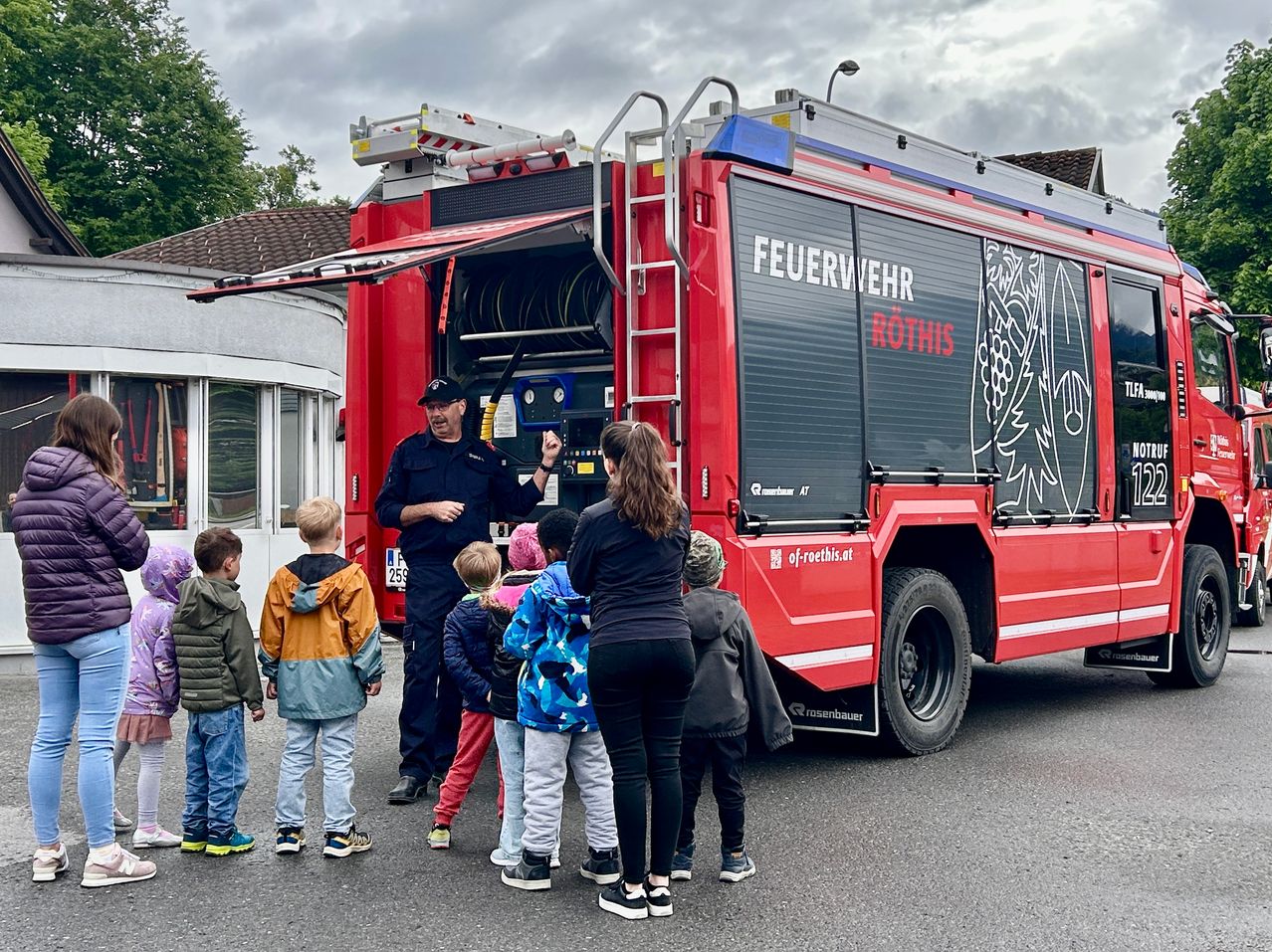 Ein Feuerwehrmann spricht vor einem roten Feuerwehrauto mit Kindern. Das Auto ist mit 'Feuerwehr Rothis' beschriftet.