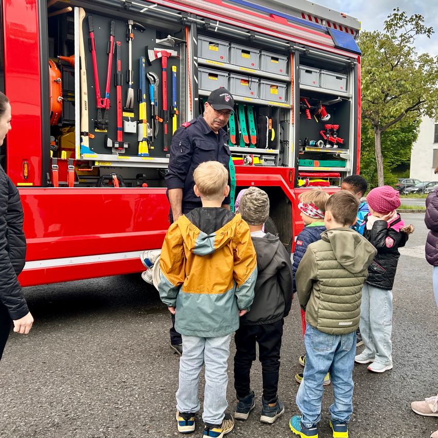 Ein Feuerwehrmann steht neben einem Feuerwehrauto und zeigt einer Gruppe von Kindern dessen Werkzeuge.