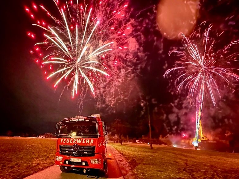 Ein roter Feuerwehrwagen fährt auf einer Straße, während in der Nacht Feuerwerk am Himmel explodiert.