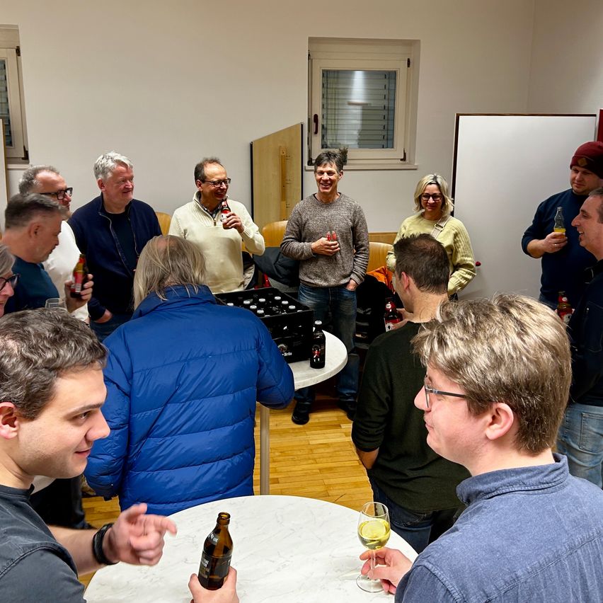 Eine Gruppe von Menschen versammelt sich in einem Raum, einige mit Getränken in der Hand. Ein Mann in einem gelben Pullover schaut einen Mann in einem grauen Pullover an. Ein Mann in einem blauen Mantel hält eine Bierflasche. Ein weißer Tisch hat eine Flasche und ein Glas Wein darauf.