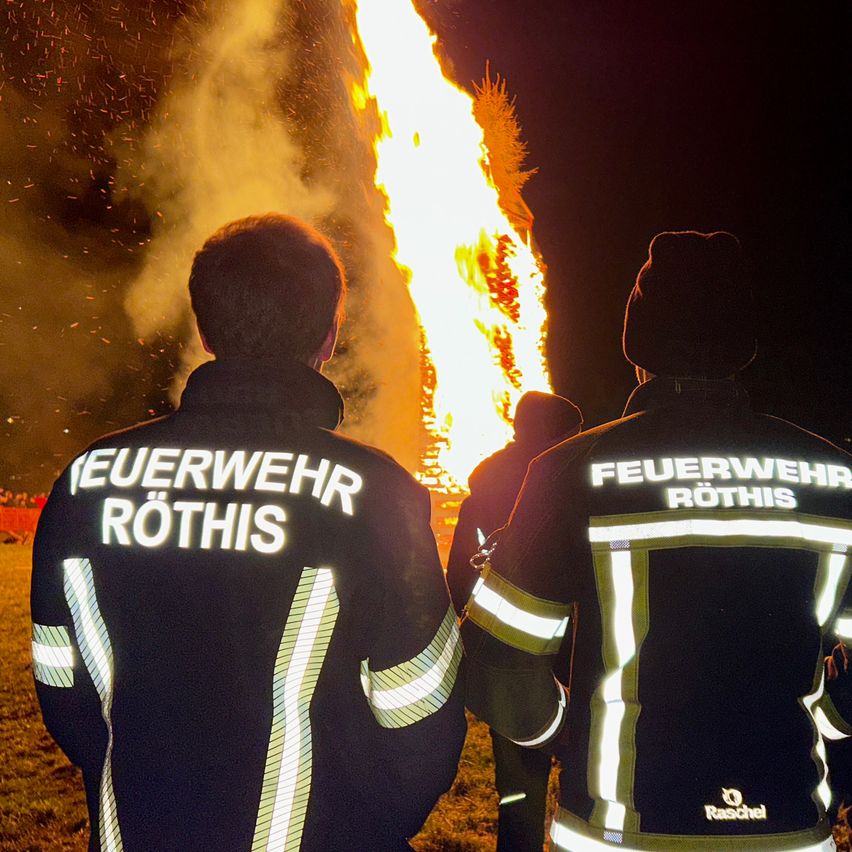 Drei Feuerwehrleute aus Rothis stehen nachts vor einem brennenden Lagerfeuer. Sie tragen reflektierende Jacken mit dem Text 'FEUERWEHR ROTHIS' auf dem Rücken.