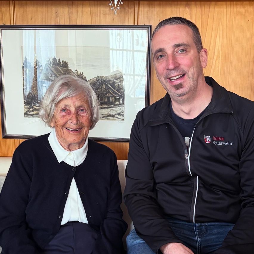 Eine lächelnde ältere Frau und ein Mann in einer Jacke mit Logo sitzen nebeneinander. Hinter ihnen ist ein Bilderrahmen mit einer Landschaft.