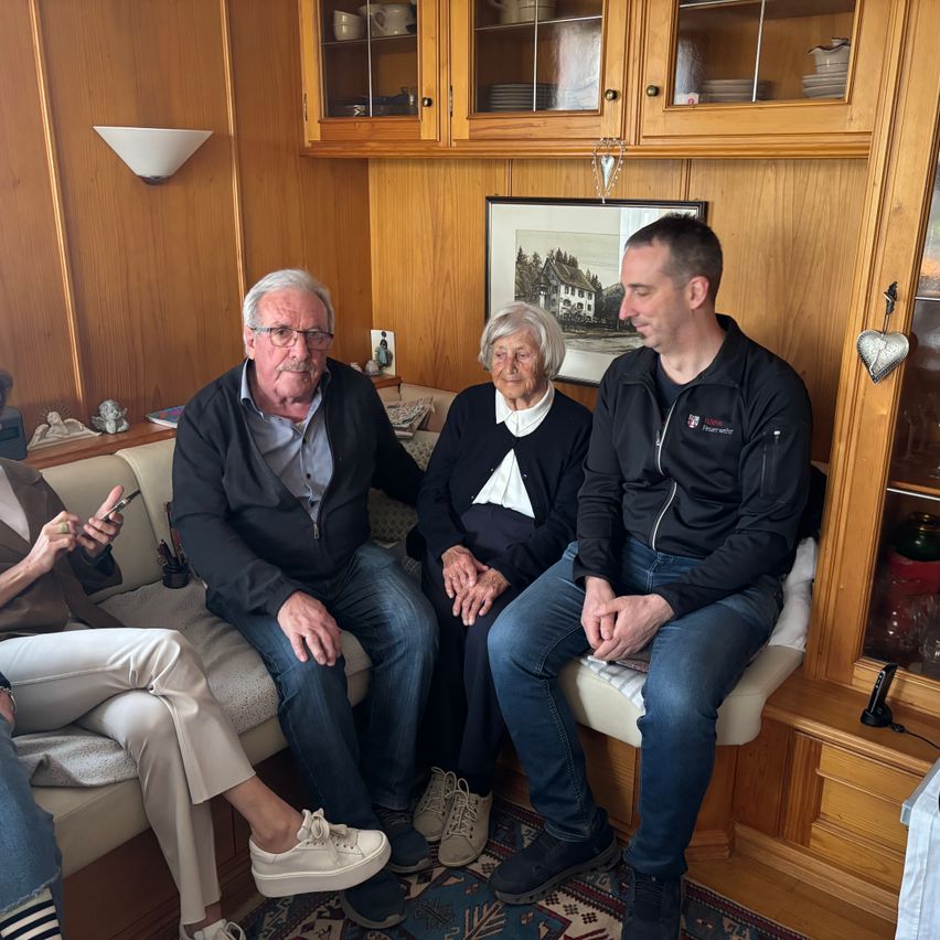 Eine Gruppe von Menschen sitzt auf einer Couch, zwei Männer und zwei Frauen, in einem hölzernen Raum mit Glasvitrinen und einem gerahmten Bild an der Wand.