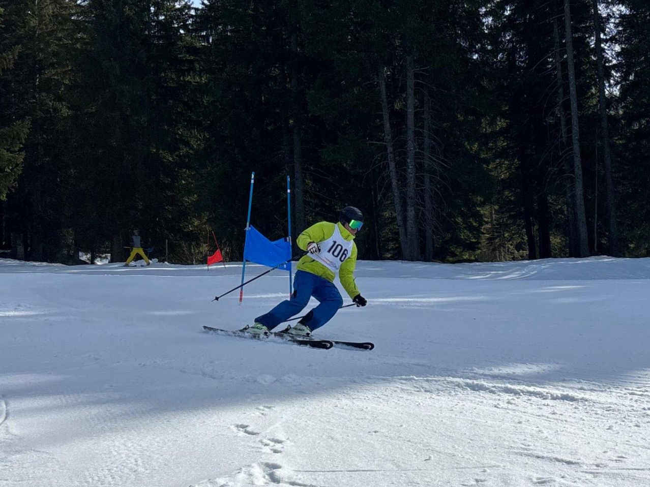 Ein Skifahrer in einem gelben Jackett mit der Nummer 106 navigiert durch einen verschneiten Parcours mit blauen Flaggen.
