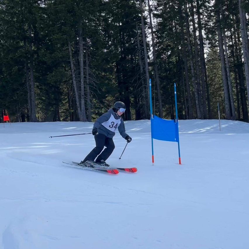 Ein Skifahrer mit der Nummer 34 gleitet einen verschneiten Hang hinunter und passiert einen blauen Marker. Im Hintergrund sind hohe Bäume und ein klarer Himmel zu sehen.