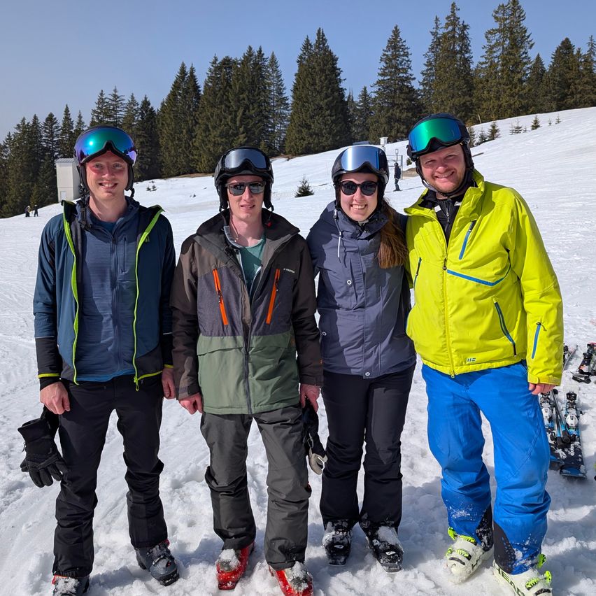 Vier Personen stehen im Schnee, gekleidet in Winterkleidung, lächeln für ein Foto. Sie tragen Helme und Schutzbrillen. Hinter ihnen ist der Schnee mit Kiefernbäumen bedeckt.