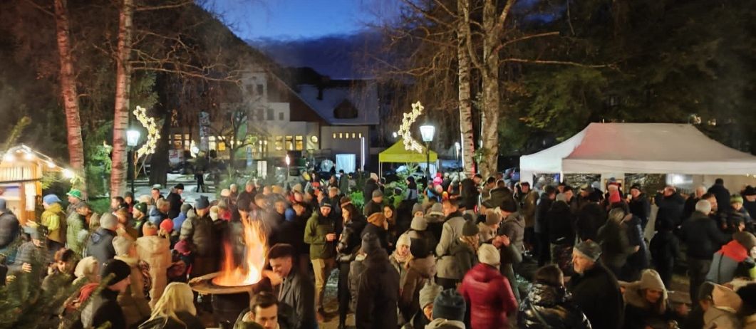 Ein nächtliches Treffen von Menschen um ein Lagerfeuer in einem Park, mit Zelten, Bäumen und einem Gebäude im Hintergrund.