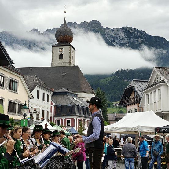 Ein Mann dirigiert eine Band auf einem Stadtplatz mit Bergen im Hintergrund. Menschen versammeln sich unter Zelten und Gebäuden.
