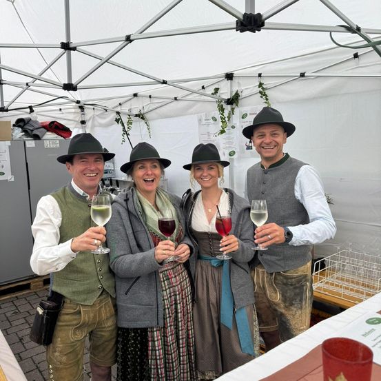 Vier Menschen in traditioneller bayerischer Kleidung posieren unter einem weißen Zelt und halten Gläser mit Getränken in den Händen. Sie lächeln und scheinen sich auf einem Festival zu befinden. Hinter ihnen sind ein weißer Lagerkasten und ein Tisch mit Gläsern zu sehen.