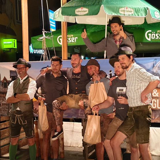 Eine Gruppe von Männern in traditioneller bayerischer Kleidung steht zusammen, einige halten Getränke und Papiertüten, einer gibt unter einem grünen Regenschirm einen Daumen hoch.