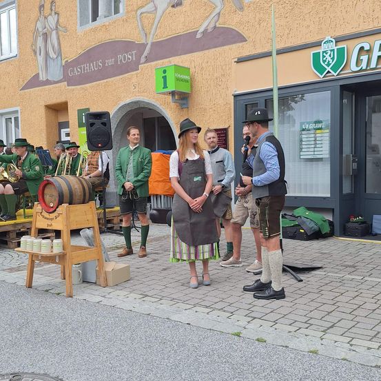 Eine Gruppe von Menschen in traditioneller deutscher Kleidung versammelt sich vor einem Gebäude. Eine Frau steht neben einem Fass, und ein Mann spricht in ein Mikrofon.