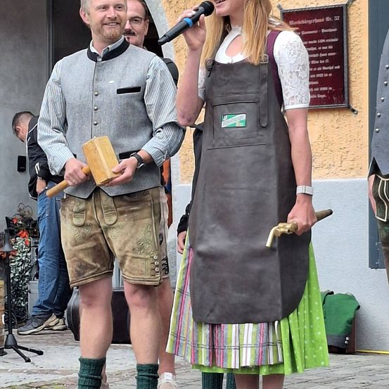 Ein Mann und eine Frau in traditioneller Kleidung stehen auf einer Kopfsteinpflasterstraße. Der Mann hält einen Holzhammer und ein Mikrofon, während die Frau einen Kupferhahn hält. Hinter ihnen ist eine Wand mit einem Schild und einer weiteren Person.