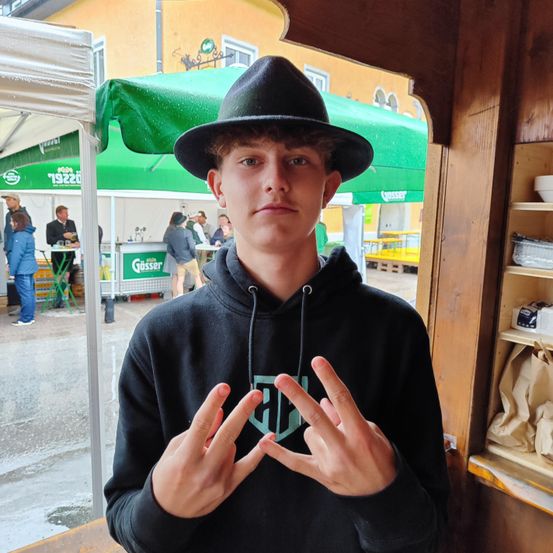 Ein junger Mann in einem schwarzen Hut und einer Kapuzenjacke steht vor einem Geschäft und macht mit seinen Fingern ein Friedenszeichen.