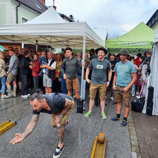 Mehrere Personen nehmen an einem Steinklopfen-Spiel teil, wobei ein Mann kurz davor steht, einen Stein eine Holzrinne hinunterzuwerfen, während andere zusehen.