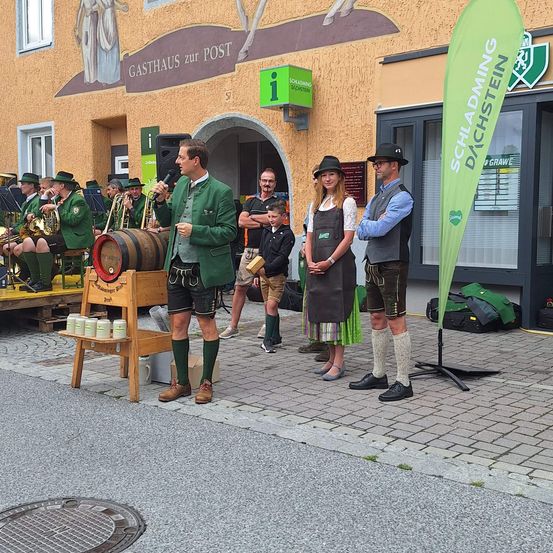 Ein Mann spricht in ein Mikrofon vor einem Fass, während eine Frau und ein anderer Mann neben ihm stehen. Sie sind vor einem Gebäude mit einem Banner. Dahinter spielen Musiker Instrumente auf einer Bühne.