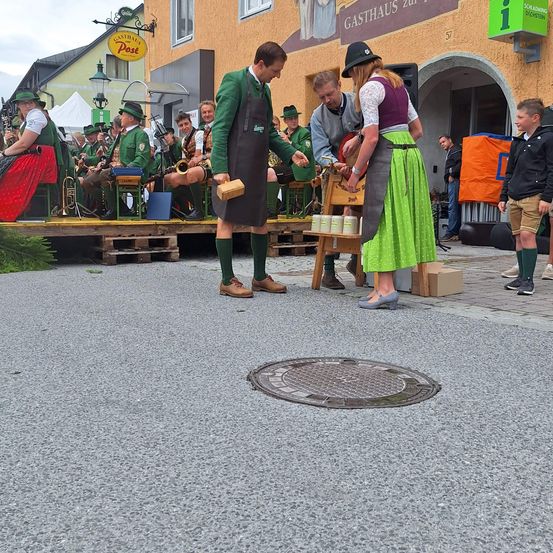 Eine Gruppe von Menschen in traditioneller bayerischer Kleidung steht auf einer Bühne. Ein Mann und eine Frau in traditioneller Kleidung stehen vor einem Tisch mit Essen. Hinter ihnen spielt ein Mann ein Instrument, während andere zuschauen.