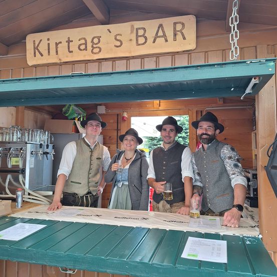 Vier Personen, gekleidet in traditioneller Kleidung, stehen hinter einer Bartheke. Ein hölzerne Schild über ihnen sagt Kirtag's Bar. Sie lächeln und posieren für ein Foto.