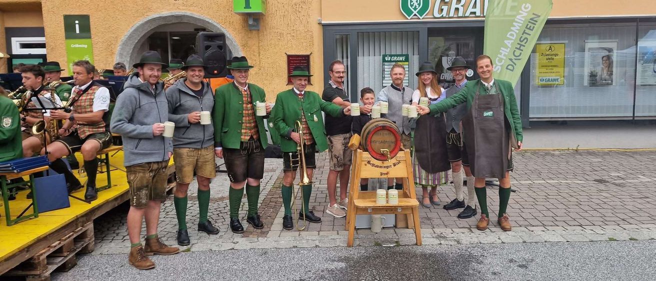 Eine Gruppe von Männern in traditionellen grünen Outfits steht vor einem Gebäude mit einem Pferdewandbild und hält Tassen und einen Fass.