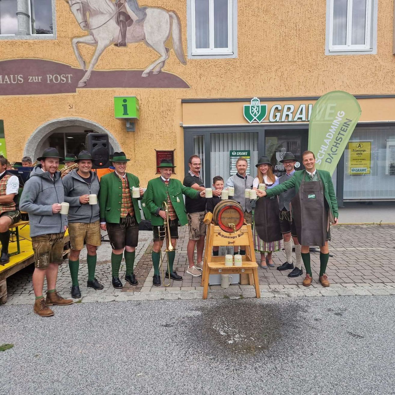 Eine Gruppe von Männern in traditioneller grüner Kleidung posiert vor einem Gebäude für ein Foto und hält Tassen. Das Gebäude hat eine Wandmalerei eines Ritters auf einem Pferd.