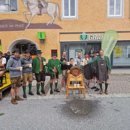 Eine Gruppe von Männern in traditioneller grüner Kleidung posiert vor einem Gebäude für ein Foto und hält Tassen. Das Gebäude hat eine Wandmalerei eines Ritters auf einem Pferd.