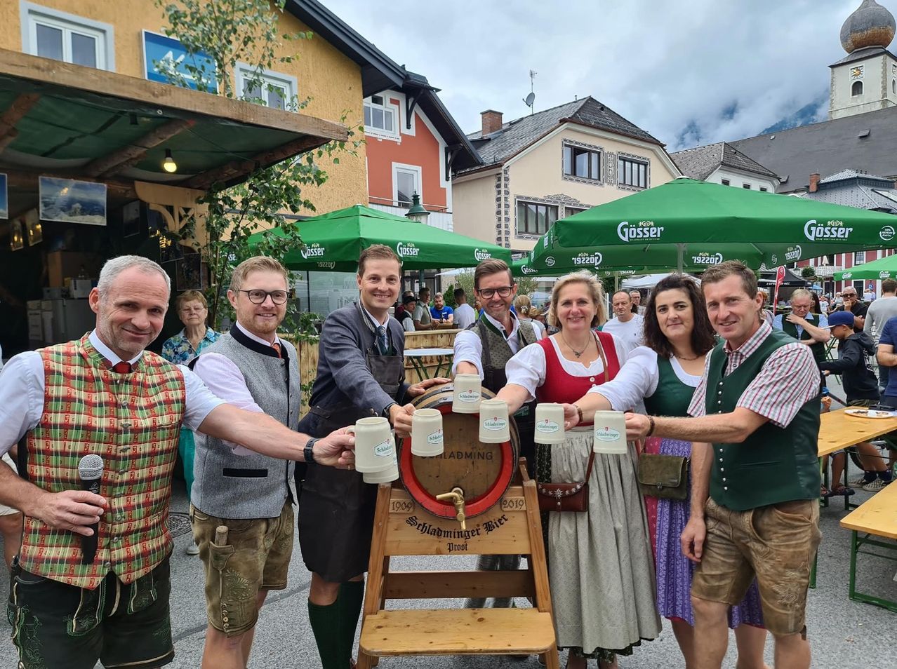 Sechs Menschen lächeln für ein Foto mit Bierkrügen in der Hand, die um ein Fass mit der Aufschrift 'Schladminger Bier' bei einem Festival stehen. Gebäude mit grünen Schirmen und Menschen in traditioneller Kleidung sind im Hintergrund zu sehen.