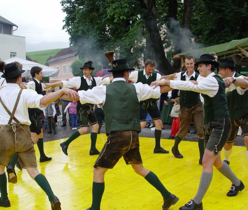 Eine Gruppe von Männern in traditioneller bayerischer Kleidung tanzt zusammen auf einem gelben Boden. Sie tragen grüne Westen und Ledershorts. Hinter ihnen stehen mehrere Bäume und ein Gebäude.