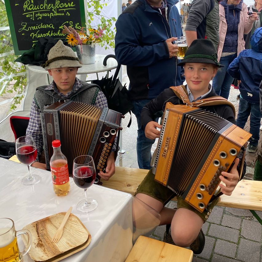 Zwei Jungen spielen Akkordeons bei einer Veranstaltung im Freien. Einer sitzt und hält sein Instrument, während der andere steht und lächelt. Weingläser, eine Flasche und ein Bierkrug stehen auf einem Tisch vor ihnen. Ein Kreidetafel mit Schrift steht hinter ihnen.